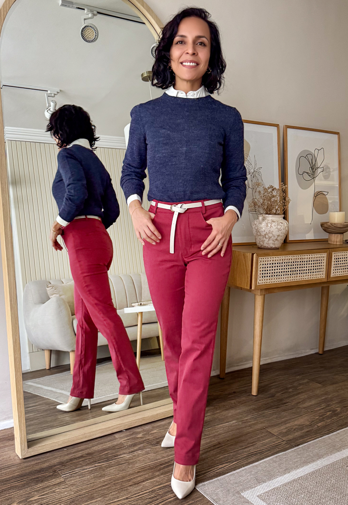 Woman in a blue sweater and pink pants standing in front of a mirror in a room with decor.