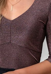 Close-up of a woman wearing a sparkling metallic plum-brown V-neck top with three-quarter sleeves and a delicate gold bar necklace.